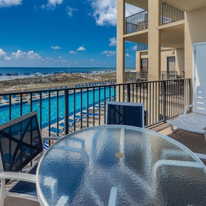 A balcony features a glass table and chairs, overlooking the sparkling pool and the beach with umbrellas in the distance