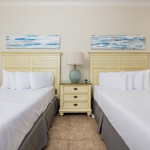 This bedroom features two comfortable full beds, light yellow cabinetry, and soothing blue artwork for a relaxing stay