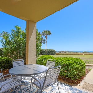 Ground floor utdoor patio area featuring a round table with chairs, surrounded by lush greenery offering a beachfront view