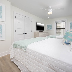 Bright guest bedroom features two queen beds with soft white linens, a TV, and coastal artwork, creating a serene atmosphere