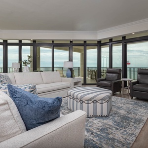 The living area features a plush sectional sofa, stylish armchairs, and stunning beach views through balcony windows