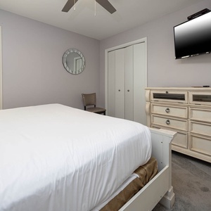 Guest bedroom features a king bed, elegant dresser, wall-mounted TV, and natural light from shuttered windows