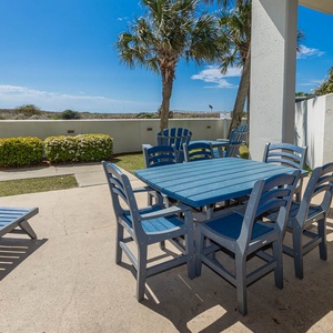 Outdoor patio area features a blue table with matching chairs, surrounded by greenery and palm trees under a sunny sky