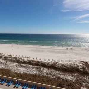 A serene view showcases the beach with white sand and gentle waves, framed by a clear blue sky and a pool area below