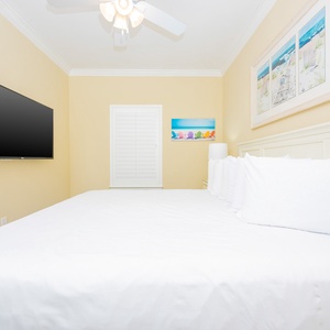 Brightly decorated guest bedroom featuring a king bed with plush white linens, a wall-mounted TV, and cheerful beach artwork