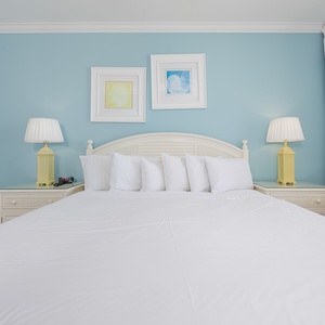 The bright master bedroom features a large king bed, complemented by cheerful yellow lamps and coastal decor