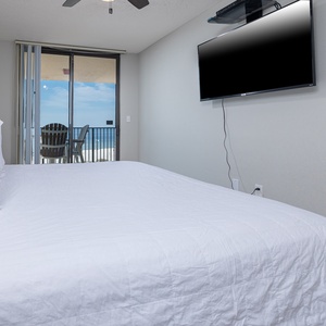 A cozy master bedroom features a king bed, flat-screen TV, and sliding doors leading to a private balcony with beach views