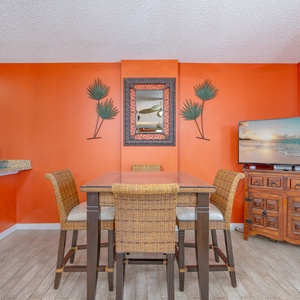 The dining area features a glass-top table with wicker chairs, a large TV, and a stunning beach view through sliding doors