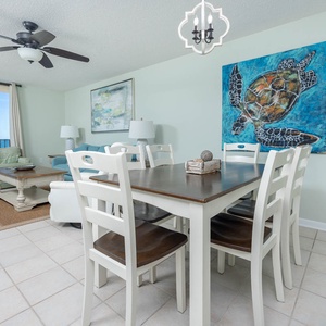 Bright dining area features a wooden table surrounded by white chairs, with a beach view visible through large windows
