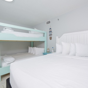 Bright guest room features a queen bed alongside a double bunk bed, with beach decor and ample white bedding for comfort