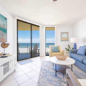 The living area features a large TV, comfortable seating, and a sliding door opening to a balcony with a beach view