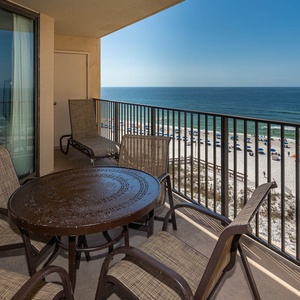 A balcony features a round table and four chairs, offering a serene view of the beach and gentle waves below