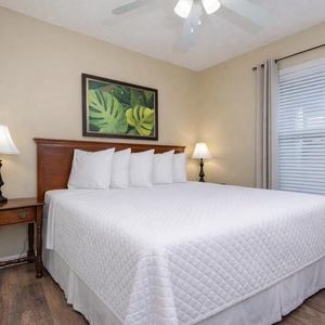 This bedroom features a large bed with crisp white linens, a tropical wall art piece, and wooden accents for a relaxing stay