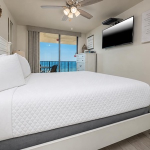Bright master bedroom featuring a plush king bed, a wall-mounted TV, and a balcony door revealing a serene beach view