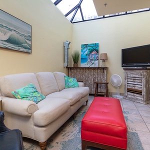 Cozy living space features a light sofa with colorful pillows, a vibrant red ottoman, and TV in the indoor master patio