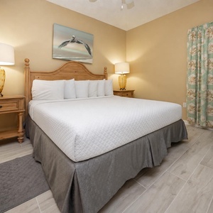 A cozy bedroom features a king bed with crisp white linens, decorative lamps, and a charming beach-themed artwork above