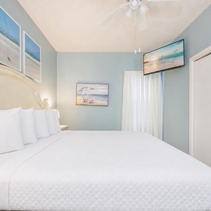 A cozy bedroom features a plush king bed, a mounted TV, and beach-themed artwork against soft gray walls