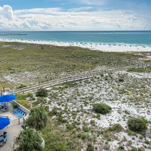View of the pool area with blue umbrellas and lounge chairs, leading to the beach with white sand and the tranquil gulf