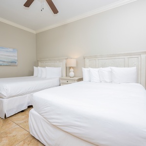 This guest bedroom features two queen beds with fresh white linens, a stylish lamp, and a calming beach-inspired painting