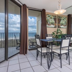 Dining area featuring a glass-top table with seating, framed tropical artwork, and a view of the beach through large windows