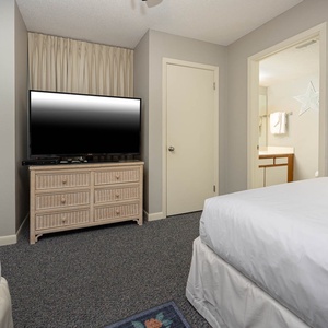 A cozy guest bedroom features two double beds, a TV on a wicker dresser, and a bathroom entrance visible in the background