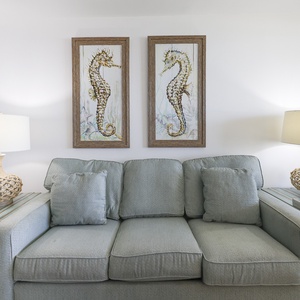 A light blue sofa with decorative pillows is complemented by beach artwork and a woven lamp on a side table