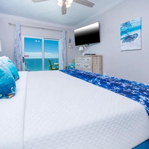 The master bedroom features a king-sized bed with vibrant blue accents and a stunning beachfront view from the sliding doors