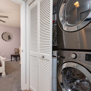 A laundry nook features a stacked washer and dryer alongside a closet door, with a glimpse of a softly colored bedroom beyond
