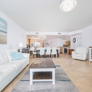 Bright living area featuring a white leather sofas with two queen sleepers, elegant chandelier, and a view into the kitchen