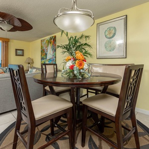 A round dining table with four chairs is set against a vibrant wall art backdrop, with a view of the beach beyond