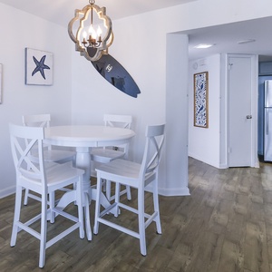 Bright dining area featuring a round white table with four chairs, coastal artwork, and a view into the kitchen
