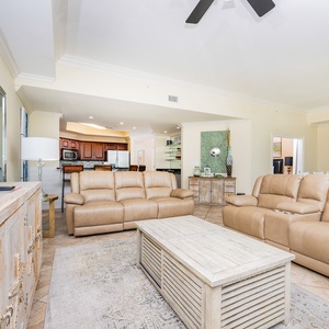 The living area features comfortable tan leather seating, a large TV, and a view into the modern kitchen with wood cabinets