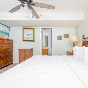 Cozy bedroom features a king bed with white linens, a wooden dresser, and a mounted TV, complemented by beach decor