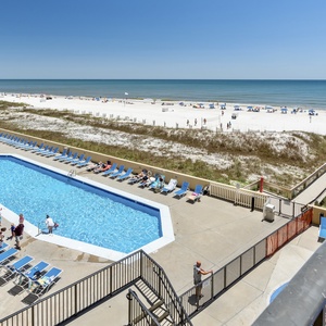 Large pool area with sun loungers, leading to a beach with white sand and colorful umbrellas in the distance