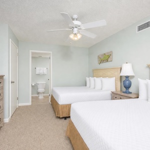 Bright guest bedroom featuring two double beds, a large TV, and a cozy dresser, with a bathroom visible in the background