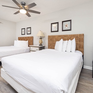 The guest room features two queen beds with woven headboards, a stylish lamp, and coastal art, creating a relaxing atmosphere