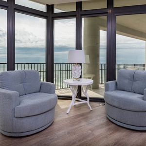 The living area features two comfortable swivel chairs, a stylish lamp, and stunning beach views through the balcony windows
