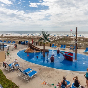 The vibrant pool area features a playful pirate ship-themed water splash zone, lounge chairs, and a scenic view of the beach