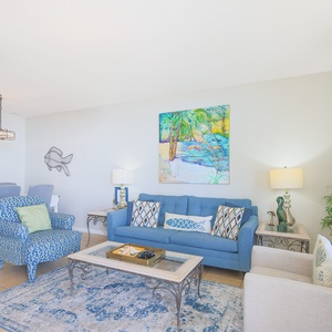 Cozy living area features a blue sofa with decorative pillows, a glass coffee table, and vibrant beach-themed artwork
