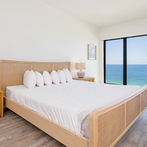 Bright and airy master bedroom featuring a king bed with plush pillows and views of the sparkling sea through large windows