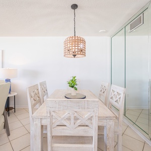 Bright dining area featuring a table surrounded by white chairs, with a beach view visible through large windows