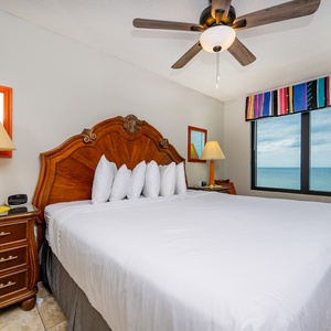This bedroom features a king bed with plush pillows, a colorful window treatment, and a stunning beachfront view