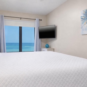 The master bedroom features a plush king bed, a TV, and stunning beachfront views through large windows