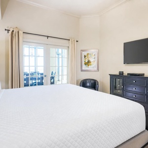 This inviting bedroom features a plush bed, a sleek TV, and large windows offering a beautiful beachfront view