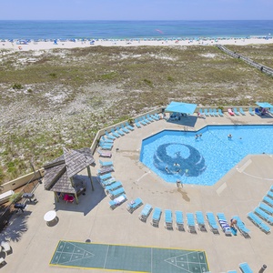 Inviting pool area with turquoise loungers, shaded gazebos, shuffleboard area, and a clear view of the beach beyond the dunes