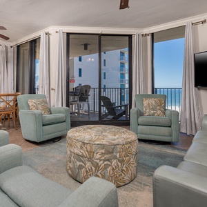Cozy seating surrounds a patterned ottoman, with sliding glass doors revealing a balcony and a glimpse of the beach beyond