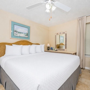 A cozy bedroom features a large king bed with white linens, a stylish wicker headboard, and a TV next to a charming dresser