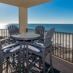 The balcony features a round table with tall chairs, offering a serene view of the tranquil beachfront and shimmering water