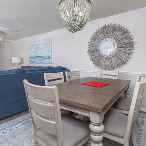 Dining area features a wooden table with six chairs, a stylish mirror, and a view of the beach through large windows
