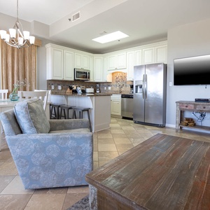 Bright living space features a comfortable blue armchair, dining table, and a modern kitchen with sleek cabinetry and a TV